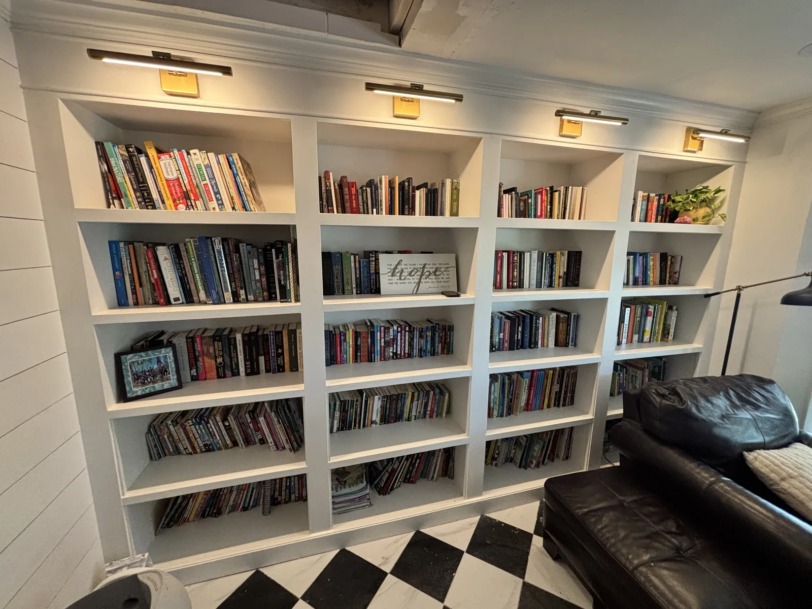 Completed custom white built-in library with floor-to-ceiling shelves, picture lights, and a checkered floor.