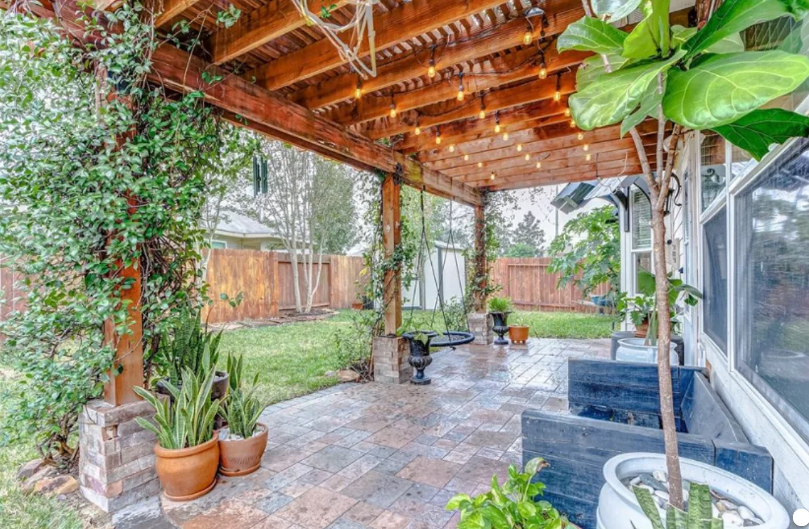 Wood pergola over a paved backyard patio with climbing plants and string lights.
